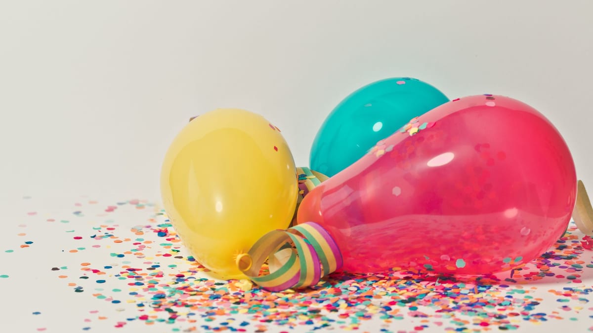 Globos amarillo, turquesa y rosa con confeti de colores esparcido sobre una superficie blanca