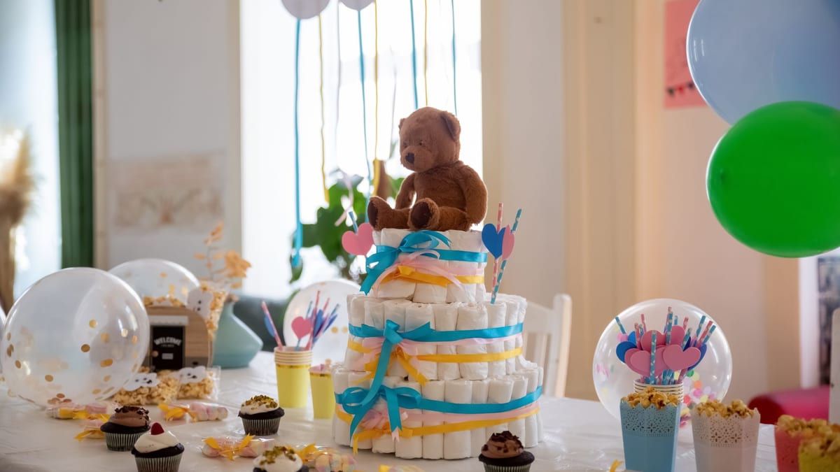 Mesa de baby shower con una tarta de pañales coronada por un osito de peluche, cupcakes y globos