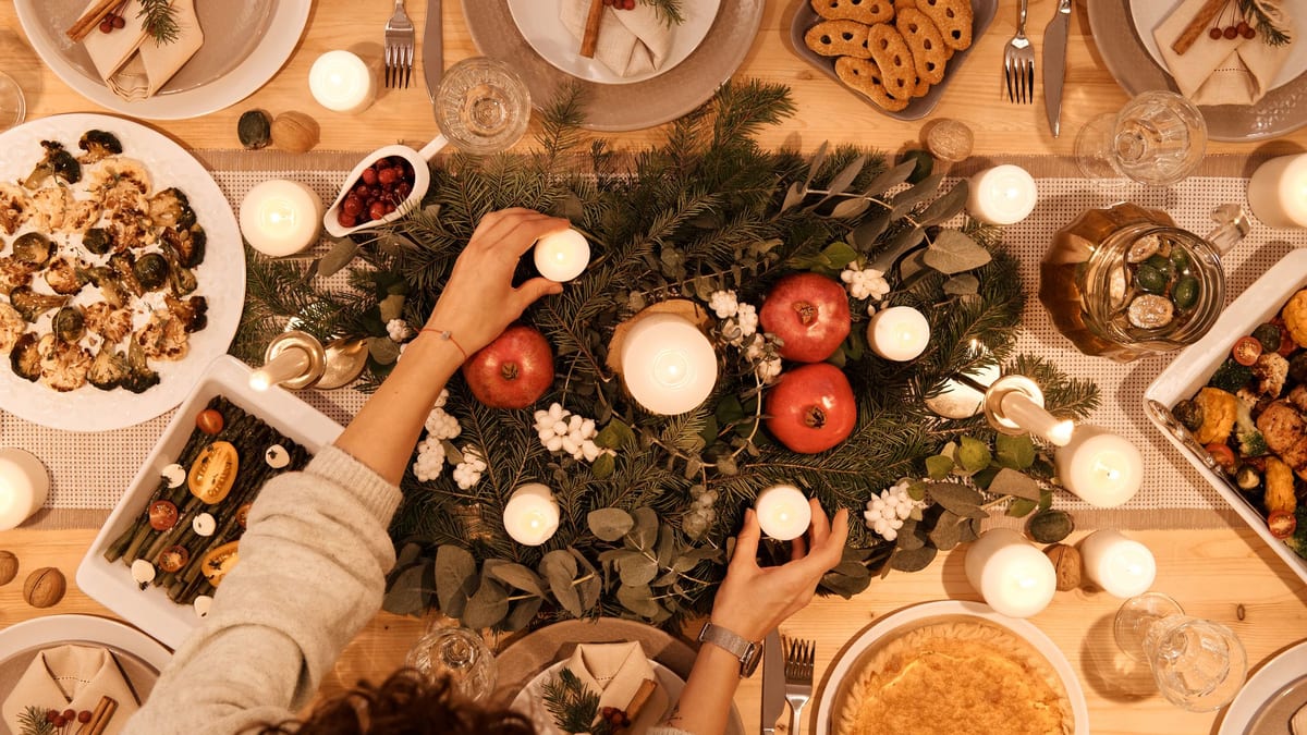 Vista cenital de una mesa de Navidad con velas, ramas de pino, granadas y platos preparados mientras alguien coloca una vela
